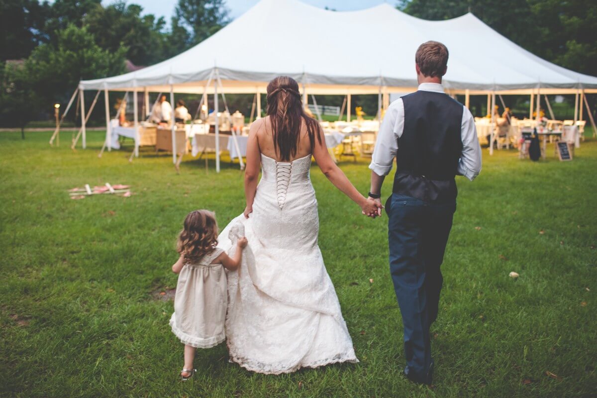 bride in wedding dress holding hands with groom in vest and jeans walking toward tent with daughter