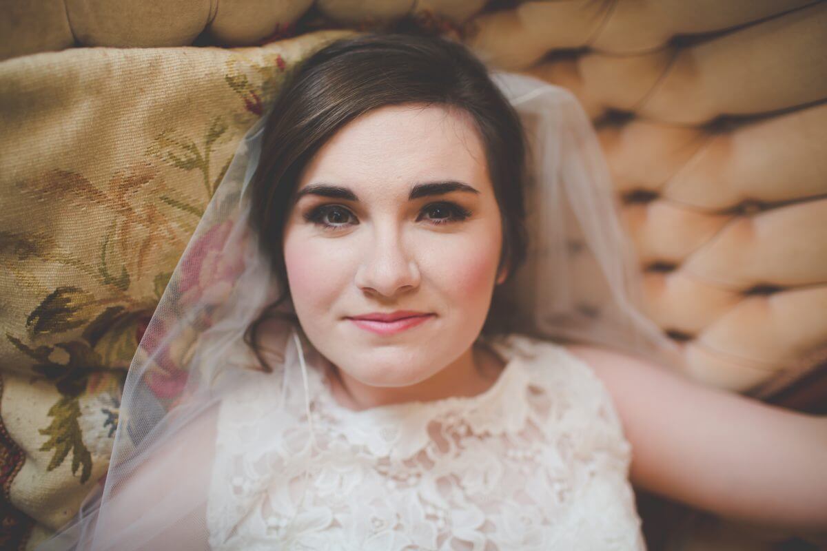 bride in dress and veil lying on vintage couch and pillow and looking up at camera