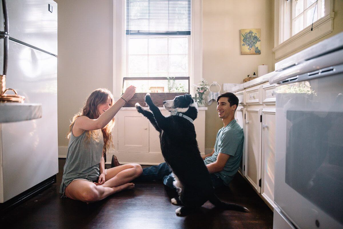 long haired woman and man sitting on kitchen floor and playing with their dog