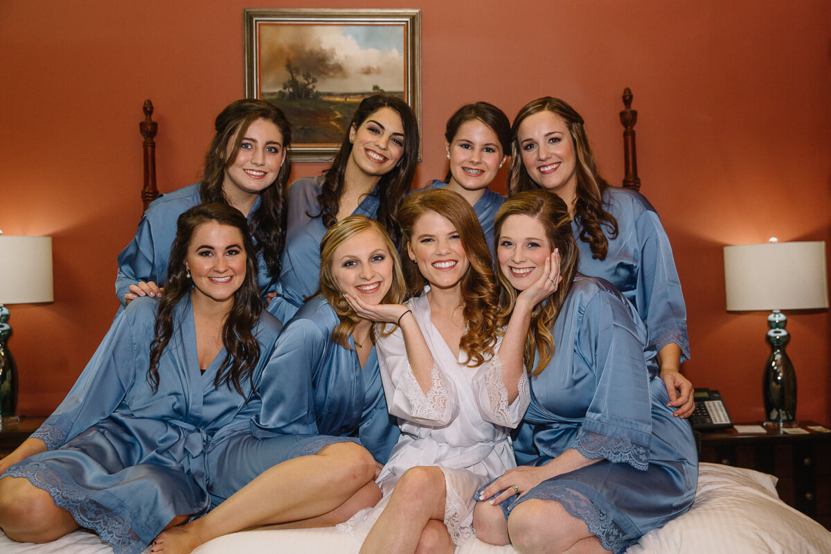 bridesmaids in blue robes sitting on bed with redheaded bride in white robe
